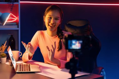 Smiling creative young beautiful Asian influencer recording on camera with positive podcast speech at night time, using laptop for presentation online at pink neon light modern studio. Stratagem. clipart