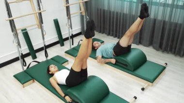 Pilates seansı sırasında iki müşteri yay namlu teçhizatı kullanarak bacak kaldırma pratiği yapıyor. Mutlu antrenör grubu karın kaslarını ve alt karın kaslarını geliştirmek için makas ya da bacak esneme pozu verir. Alışkanlık.