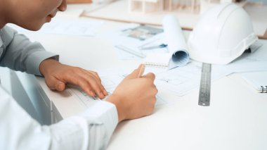 Professional architect engineer drawing blueprint and working together with young beautiful caucasian coworker on meeting table with safety helmet, house model, blueprint scatter around. Immaculate. clipart