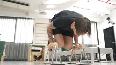 Spor salonunda vücut kontrolünü ve çekirdek gücünü arttırmak için pilates reformcusu üzerinde L-sit egzersizi yapan bir adam. Sportif adam, çekirdek, kol ve omuz güçlendirmek için yenilikçiyi sıkıştırıyor. Alışkanlık.