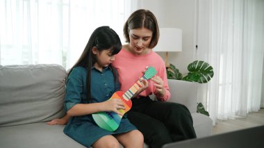 Kafkasyalı bir anne evde akustik müzik öğretirken ukulele çalan mutlu kız. Sevimli çocuk enstrüman öğreniyor. Kafkasyalı mutlu anne ve kız birlikte vakit geçiriyorlar. Pedagoji.
