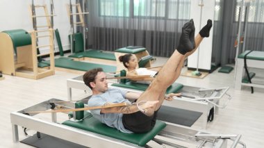 Bir grup müşteri, yenilikçi bir makinede bacak yükseltme işlemi yapıyor. Pilates partneri reformcu pozu veriyor ve egzersiz sırasında dengeyi geliştirmek ve koordinasyonu arttırmak için kayış takıyor. Alışkanlık.