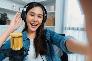 Sunucu kanalı genç ve güzel Asyalı kız akıllı telefonuyla selfie çekiyor. Modern stüdyo ofis konseptinde kulaklık takıyor. Sahne arkası yayıncısı sosyal medyada canlı yayın yapıyor. Taktik.