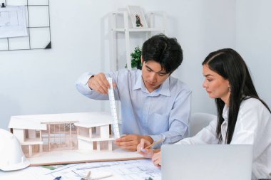 Profesyonel Asyalı erkek mimar ev modelinin uzunluğunu ölçmek için cetvel kullanırken, beyaz tenli genç meslektaşı ev modeliyle toplantı masasındaki verileri analiz etmek için dizüstü bilgisayar kullanıyor. Tertemiz..