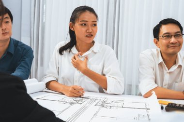 Mimari proje üzerinde birlikte çalışan çeşitli inşaat mühendisi ve müvekkil grupları, inşaat planını gözden geçirip toplantı masasında plan oluşturuyorlar. Tedbirli