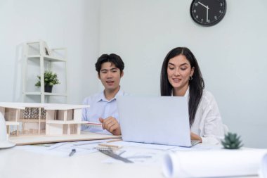 Profesyonel Asyalı erkek mimar ev modelinin uzunluğunu ölçmek için cetvel kullanırken, beyaz tenli genç meslektaşı ev modeliyle toplantı masasındaki verileri analiz etmek için dizüstü bilgisayar kullanıyor. Tertemiz..