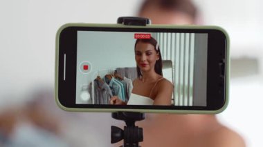 Genç sosyal medyum kadın moda videosu yapıyor. Blogcu kameraya selfie çubuğu ve ışık halkası kullanarak gülümserken, aynı zamanda ikna edici çevrimiçi kıyafetlerin izleyicilere veya takipçilere vlog satmasını sağlıyor. Düzenle