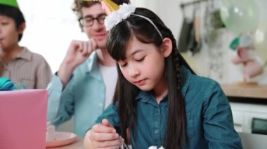 Asyalı bir kız doğum günü pastası yerken, beyaz bir anne aile üyesine hediye olarak kaşık veriyor. Aile üyesi balonla süslenmiş mutlu kız doğum gününü kutlamak için toplandı. Pedagoji.