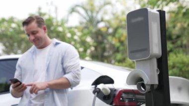 Adam doğadaki EV şarj istasyonunda elektriği ödemek için akıllı telefonu kullanıyor. Modern çevre ve EV araçlı sürdürülebilir otomobil taşıma yaşam tarzı. Titiz