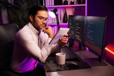 Akıllı bilişim geliştiricisi bilgisayar ekranında yazılım geliştirme kodlaması üzerinde çalışıyor ve en yeni uygulama projesinin notlarını okuyor, program güncellemesi düşünüyor neon ışıklı siber ofiste online web sitesi verileri. Tahmin et..