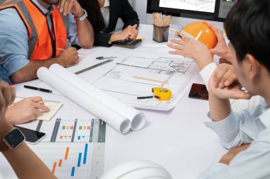 Mimari proje üzerinde birlikte çalışan çeşitli inşaat mühendisi ve müvekkil grupları, inşaat planını gözden geçirip toplantı masasında plan oluşturuyorlar. Tedbirli