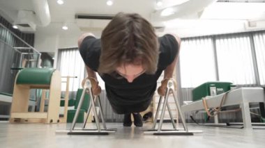 Müşteri, bir spor salonunda üst vücut gücünü, dengeyi ve çekirdek bağını geliştirmek için pilates egzersizi sırasında tutamaklarını kullanarak şınav çekiyor. Adam egzersiz yapıyor ve şınav çekiyor. Alışkanlık.