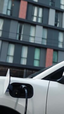 Takım elbiseli, hırslı bir işadamı elektrik kablosu ve yenilenebilir enerji enerjili bir elektrikli araçla şarj istasyonunda dururken elektrikli aracına yaslanıyor..