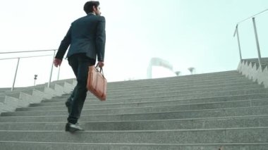 Akıllı bir iş adamının elinde çantayla merdivenlerden yukarı çıkışının en üst görüntüsü. Profesyonel proje yöneticisi merdivenlerden çıkıp işyerine gidiyor. Yeteneklerini arttırmak, terfi almak, seyahat etmek. Sevinçli..