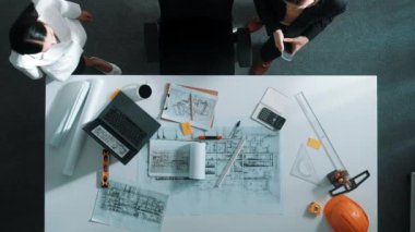 Planlara bakan, proje planı ve güvenlik kaskıyla toplantı masasında oturan mühendis ile konuşan yetenekli yöneticinin üst görüşü. Mimarların hava görüntüsü belgeyi tutar ve yürür. Hizalama.