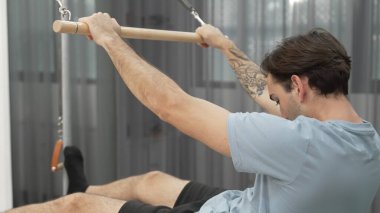 Man performing seated core exercise on pilates trapeze holding bar to strengthen arms and core muscles at gym. Trainer doing chest press on the cadillac for strengthening chest, shoulder. Habituate. clipart