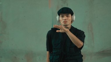 Enerjik hipster şehir caddesinde kulaklıktan hip-hop müziğiyle dans ediyor. Sokak dansı performansı çalışırken kameraya bakan break dansçı. Geçmişi. Açık hava sporu 2024. Hiphop.