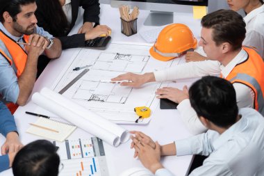 Mimari proje üzerinde birlikte çalışan çeşitli inşaat mühendisi ve müvekkil grupları, inşaat planını gözden geçirip toplantı masasında plan oluşturuyorlar. Tedbirli