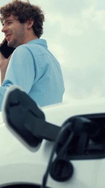 Takım elbiseli, hırslı bir işadamı elektrik kablosu ve yenilenebilir enerji enerjili bir elektrikli araçla şarj istasyonunda dururken elektrikli aracına yaslanıyor..