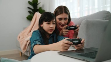 Annem dizüstü bilgisayar kullanarak kanepeye uzanırken, mutlu kız oyun oynuyor ve oturuyor. Beyaz bir ebeveyn ve çekici bir kız, elinde joystick ile birlikte vakit geçirir ve maç kazanmaya odaklanırlar. Pedagoji.