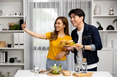 Gülümseyen sunucu şefler akıllı telefonla selfie çekerek sosyal medya kanalından canlı yayında yemek programında çift büyük hamburger yapıyorlar. İnfobahn.