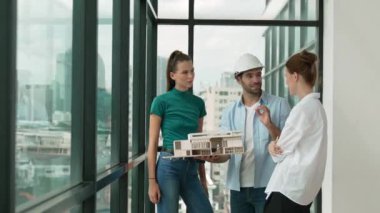 Proje yöneticisi ev modelini elinde tutarken, mimar mühendis takımı da ev tasarımı veya bina inşaatı hakkında fikir alışverişinde bulunuyor. Şehrin penceresinin yanında iç mimar duruyor. İzleme