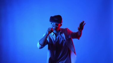 Mavi arka planda VR gözlüklerle dans edip şarkı söylerken mutlu dansçılar. Günlük kıyafetleri olan çekici bir adam enerjik hareketler yapar ve teknolojiyi kullanarak şarkı söyler. Aygıt.