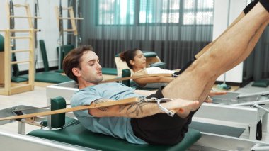 Bir grup müşteri, yenilikçi bir makinede bacak yükseltme işlemi yapıyor. Pilates partneri reformcu pozu veriyor ve egzersiz sırasında dengeyi geliştirmek ve koordinasyonu arttırmak için kayış takıyor. Alışkanlık.
