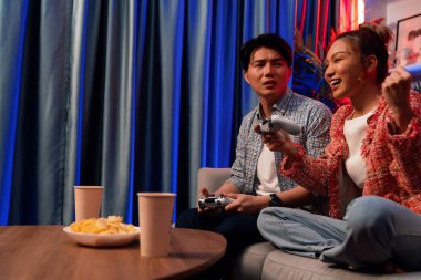 Mutlu çiftin oturduğu kanepe joysticklerle video oyunu oynuyor, meydan okuyan gülümseyen kadın, evdeki modern neon ışıklı oturma odasındaki mücadeleyi kazanmak için yumruk kaldırıyor. İnfobahn.