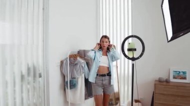 Genç sosyal medyum kadın moda videosu yapıyor. Blogcu kameraya selfie çubuğu ve ışık halkası kullanarak gülümserken, aynı zamanda ikna edici çevrimiçi kıyafetlerin izleyicilere veya takipçilere vlog satmasını sağlıyor. Düzenle