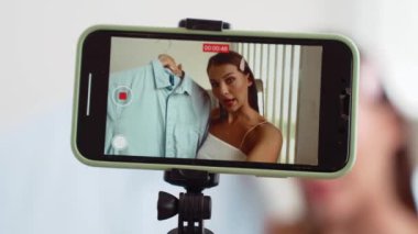 Genç sosyal medyum kadın moda videosu yapıyor. Blogcu kameraya selfie çubuğu ve ışık halkası kullanarak gülümserken, aynı zamanda ikna edici çevrimiçi kıyafetlerin izleyicilere veya takipçilere vlog satmasını sağlıyor. Düzenle