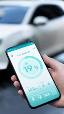 Akıllı telefon ekran bataryasını elinde tutan iş adamı akıllı EV mobil uygulama ile otomobil parkındaki şarj istasyonundan elektrik şarj eden çevre dostu EV otomobil. İnceleyin