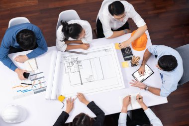 Mimari proje üzerinde birlikte çalışan, inşaat planını gözden geçiren ve toplantı masasında plan oluşturan çeşitli inşaat mühendisi ve müşteri gruplarından oluşan üst düzey bir pankart. Tedbirli