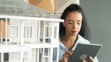 Genç, güzel İspanyol mimar mühendis modern ofisteki ev modeli yapısını kontrol ederken tablet kullanarak bina inşaatını çiziyor. Yaratıcı iş inşaat konsepti. Yönlendirici.