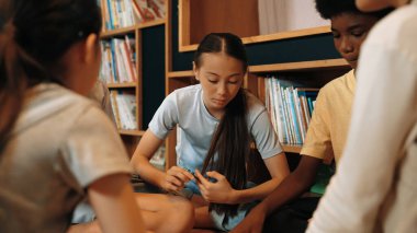 Group of diverse children sitting in circle while reading a book at library. Highschool girl sharing a magazine while pointing at interested topic and talking with multicultural friends. Edification. clipart