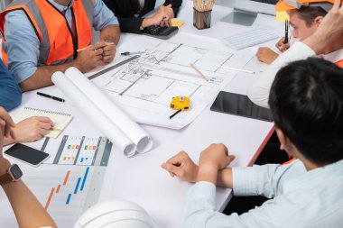 Mimari proje üzerinde birlikte çalışan çeşitli inşaat mühendisi ve müvekkil grupları, inşaat planını gözden geçirip toplantı masasında plan oluşturuyorlar. Tedbirli