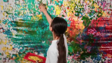 Genç ve güzel bir kızın arka görünüşü renkli duvarı boyamak için elini kaldırır. Çekici bir lise öğrencisi renk paletini tutarken duvarı el iziyle boyuyor. Yaratıcı aktivite. Öğretim