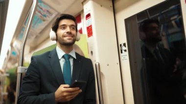 Akıllı yönetici trende dururken kulaklık takar ve müzik dinler. Profesyonel iş adamı, şarkıları dinlerken ve şarkı seçerken bölüme yaslanıyor. İş konsepti. Sevinçli..
