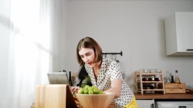 Kafkasyalı anne, modern mutfakta dizüstü bilgisayarda yemek tarifine bakarken salata yapmak için yemek hazırlıyor. Yetenekli ev bekçisi taze sebze kullanarak kahvaltı hazırlıyor. Pedagoji.