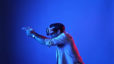 Meta-World 'de atış oyunu oynarken VR kulaklık takan profesyonel bir oyuncu. Erkek model, sanal gerçeklik gözlüğü kullanarak mavi arka planda dururken silah hareketi yapıyor. Aygıt.