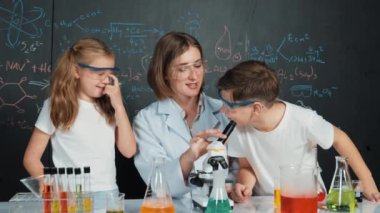 Öğretmen tavsiye verirken beyaz çocuk kimyasal sıvıyı karıştırıyor. Profesyonel eğitmen labaratuar kıyafeti giymiş, renkli solüsyonla dolu deney kabıyla sofrada çeşitli öğrencileri arıyor. Etkinlik.
