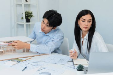 Profesyonel Asyalı erkek mimar ev modelinin uzunluğunu ölçmek için cetvel kullanırken, beyaz tenli genç meslektaşı ev modeliyle toplantı masasındaki verileri analiz etmek için dizüstü bilgisayar kullanıyor. Tertemiz..