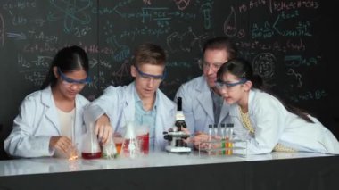 Çok kültürlü bir öğrenci mikroskop altında incelerken güzel bir kız da numuneyi inceliyor. BTMM sınıfında karatahta bilim teorisi deneyi yapan liseli çocuklara bakan öğretmen. Öğretim