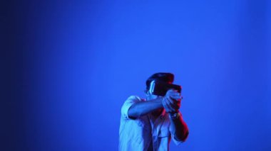 Meta-World 'de atış oyunu oynarken VR kulaklık takan profesyonel bir oyuncu. Erkek model, sanal gerçeklik gözlüğü kullanarak mavi arka planda dururken silah hareketi yapıyor. Aygıt.