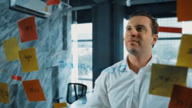 Akıllı iş adamı cam tahtanın önünde bir proje başlatmayı düşünüyor. Gülümseyen erkek proje yöneticisi yaratıcılık pazarlama stratejisini gösteren renkli yapışkan notlara bakıyor. Yönlendirici.