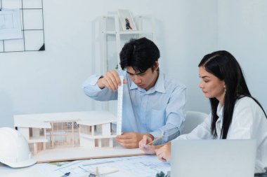 Profesyonel Asyalı erkek mimar ev modelinin uzunluğunu ölçmek için cetvel kullanırken, beyaz tenli genç meslektaşı ev modeliyle toplantı masasındaki verileri analiz etmek için dizüstü bilgisayar kullanıyor. Tertemiz..