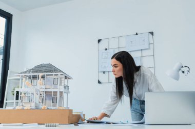 Genç, beyaz, güzel bir mimarın portresi toplantı masasına yerleştirilen ev modeli, plan ve dizüstü bilgisayarın yapısını karşılaştırmaya odaklanıyor. Şah, teftiş, tahmin konsepti. Tertemiz..