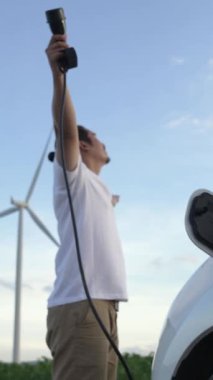 Elektrikli arabasıyla ilerici bir adam, EV arabası gelecekteki sürdürülebilir enerji kavramı olarak rüzgar türbiniyle yeşil alandaki enerji şarj etme istasyonundan enerji yüklüyor. Enerji üreteci olan elektrikli bir araç..