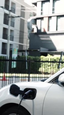 Takım elbiseli, hırslı bir işadamı elektrik kablosu ve yenilenebilir enerji enerjili bir elektrikli araçla şarj istasyonunda dururken elektrikli aracına yaslanıyor..