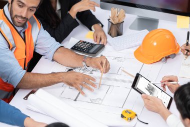 Mimari proje üzerinde birlikte çalışan çeşitli inşaat mühendisi ve müvekkil grupları, inşaat planını gözden geçirip toplantı masasında plan oluşturuyorlar. Tedbirli